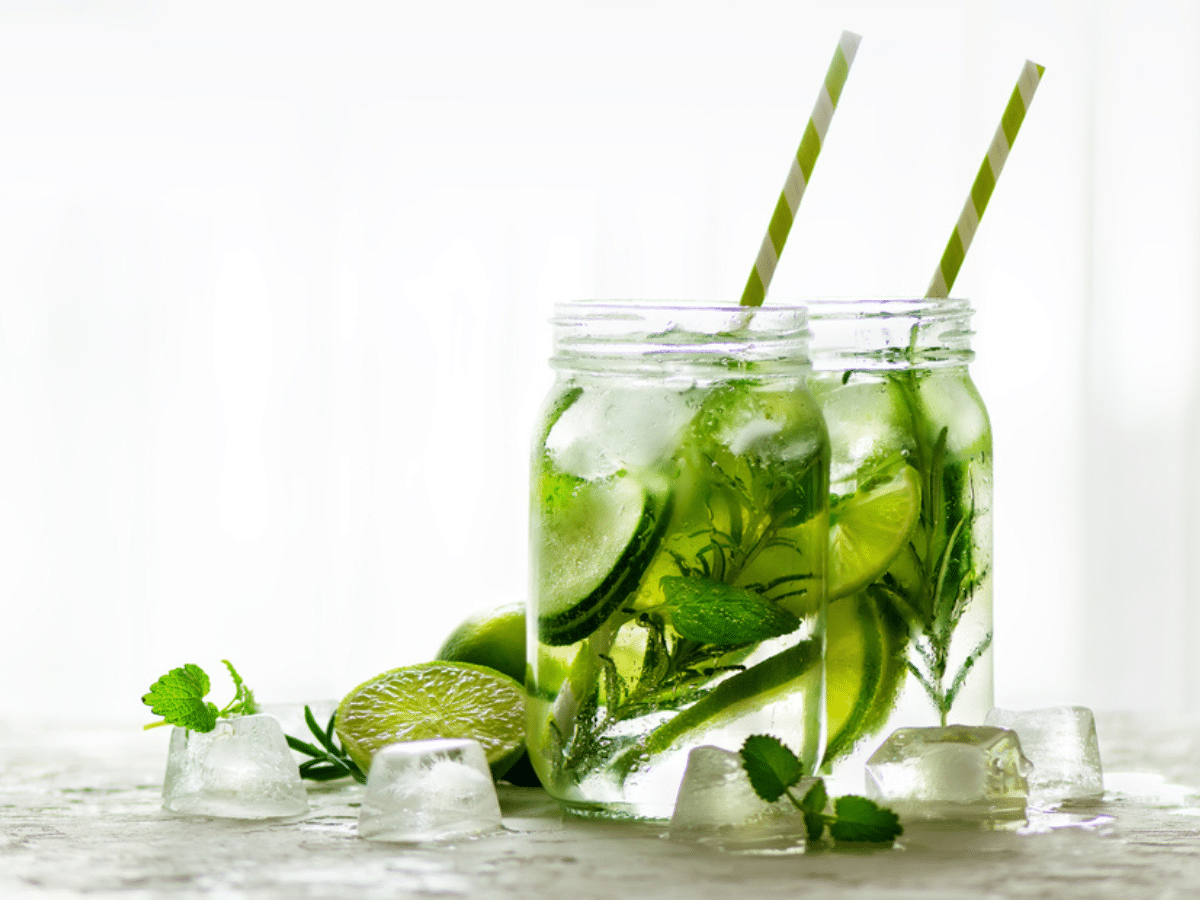 Zwei Gläser sind mit Wasser gefüllt. Darin sind Gurken- sowie Limettenscheiben, Rosmarinzweige, Eiswürfel und Minze enthalten. Die Gläser stehen auf einem Tisch.